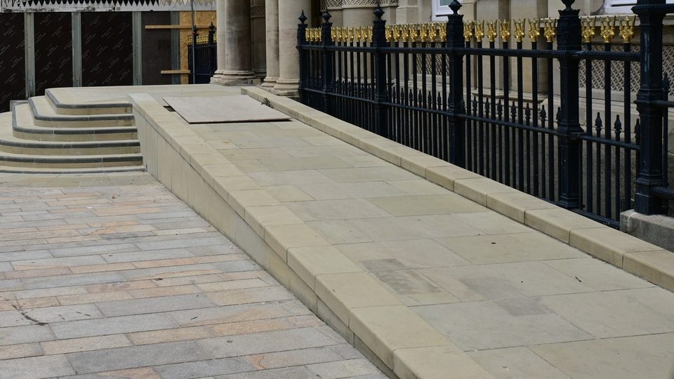 A gently sloping stone ramp running beside a short flight of steps and black iron railings at a public building
