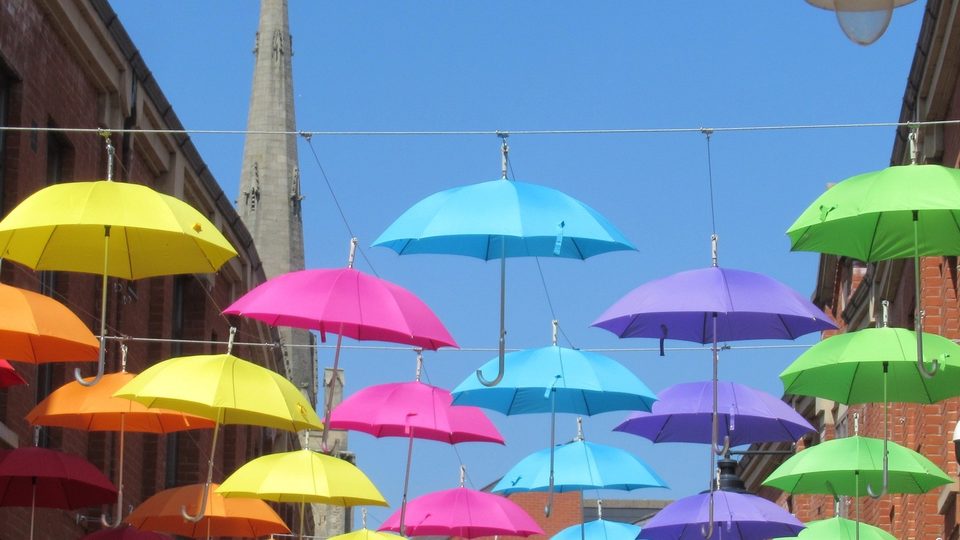 Rows of bright yellow, pink, blue, purple and green umbrellas suspended above a street against a clear blue sky