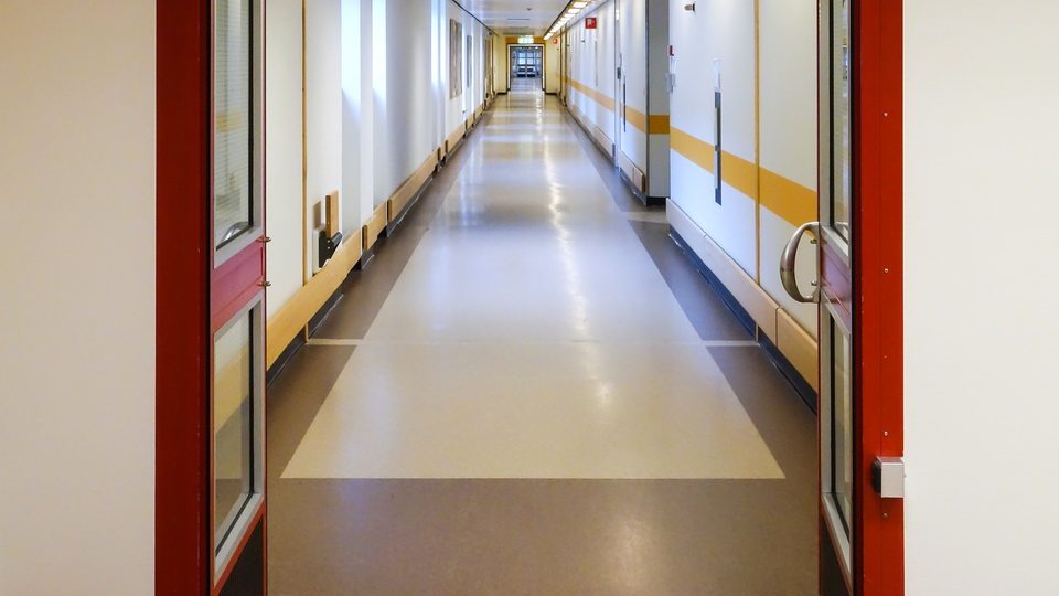 A long hospital corridor seen through an open pair of red-framed doors, its floor polished and its walls striped in yellow