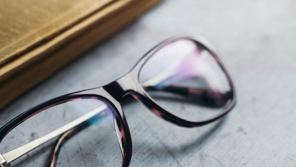 A pair of reading glasses resting on a grey surface beside the corner of an old cloth-bound book