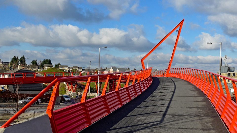 A footbridge with bright red railings curving towards a tall red mast under a blue sky with white cloud