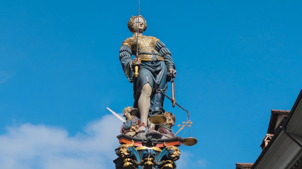 The painted statue of Justice in Bern, blindfolded and holding a sword and scales, against a bright blue sky