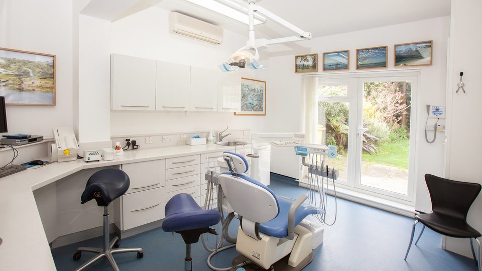 A bright, white dental surgery with a blue treatment chair and a garden view through glass doors
