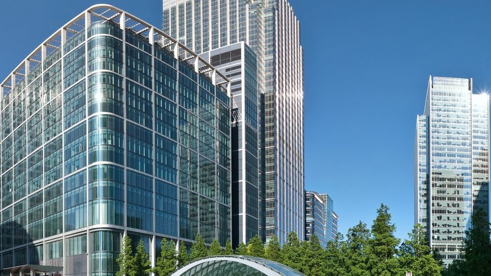 The towers of Canary Wharf, where the GPhC has its office, rising above green trees under a deep blue sky
