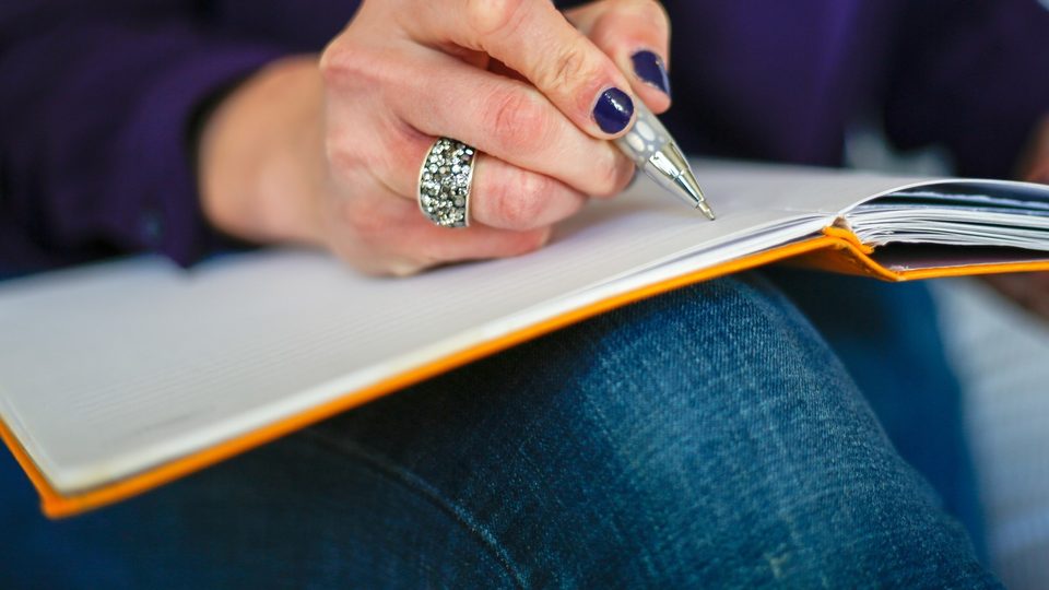 A hand with dark blue nail varnish writing in an orange-edged notebook resting on a knee