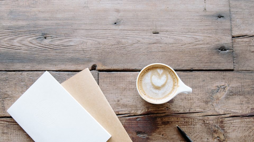 A white notebook, a brown notebook and a cup of coffee with a heart in the foam on a weathered wooden table
