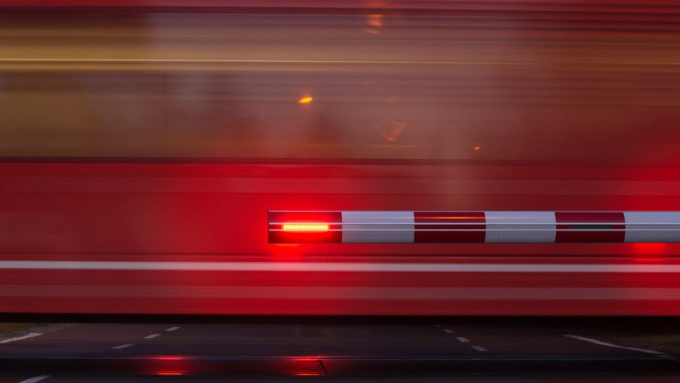 A lowered red and white level crossing barrier with its red lamp glowing, closing the road in front of a blurred red train